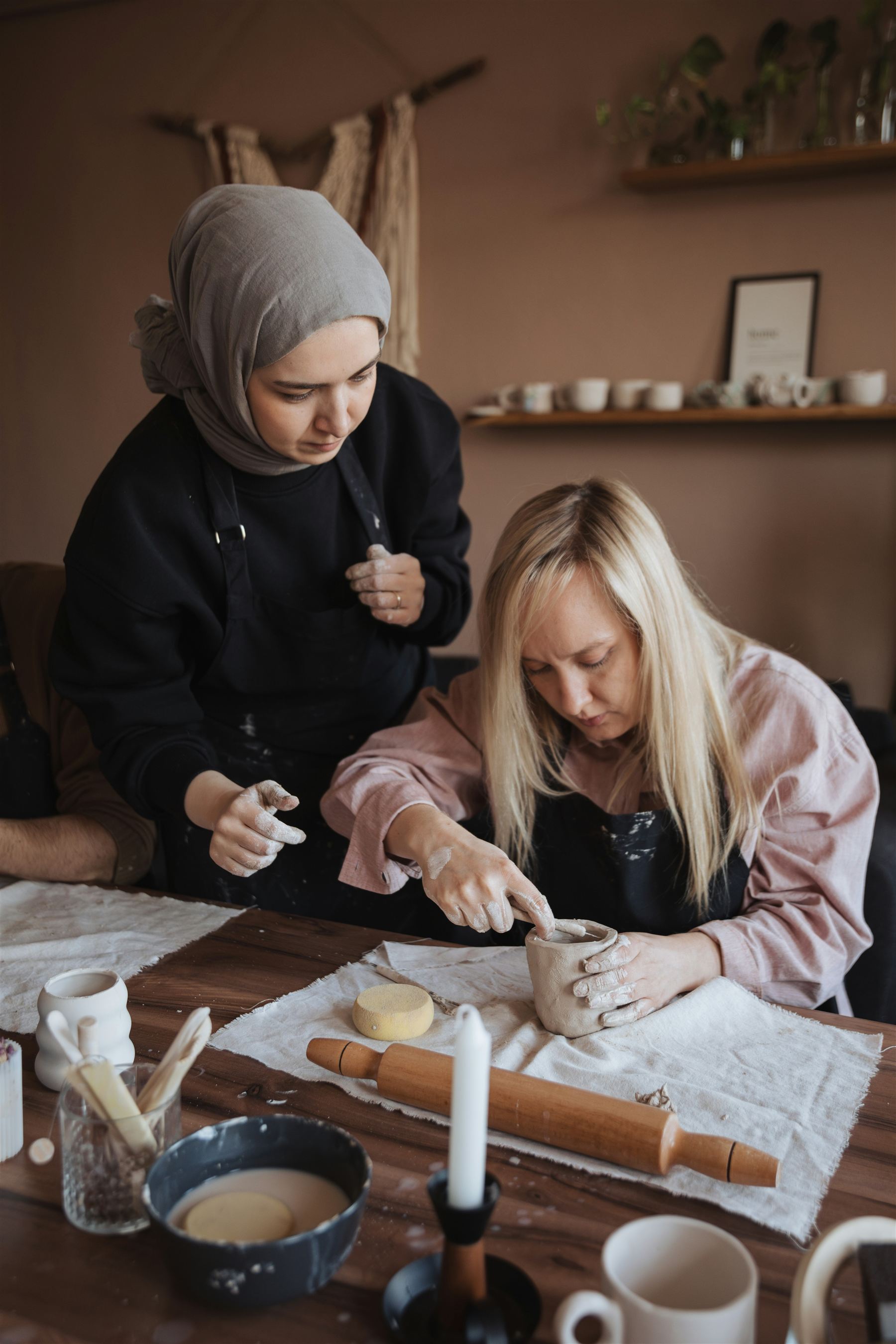 Two women sitting together at a table, one wearing a hijab and guiding the other's hands as they shape a small clay cup.