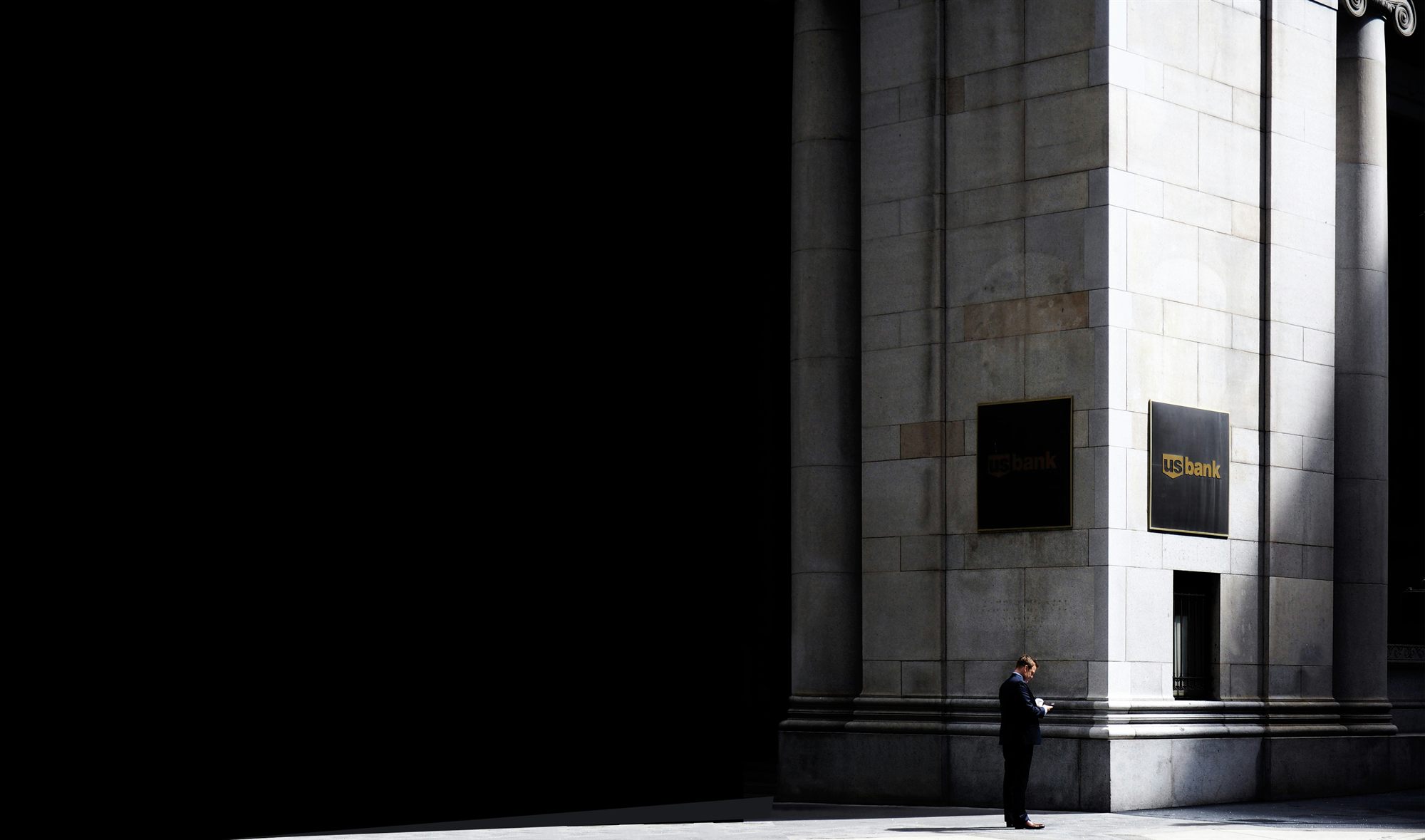 A man in a suit checking his phone in the shadow of a large stone bank building.