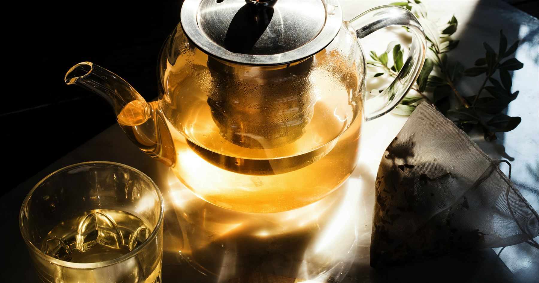 A glass teapot pouring tea into a glass cup on a dark table, with loose tea leaves and greenery nearby, lit in warm golden light.