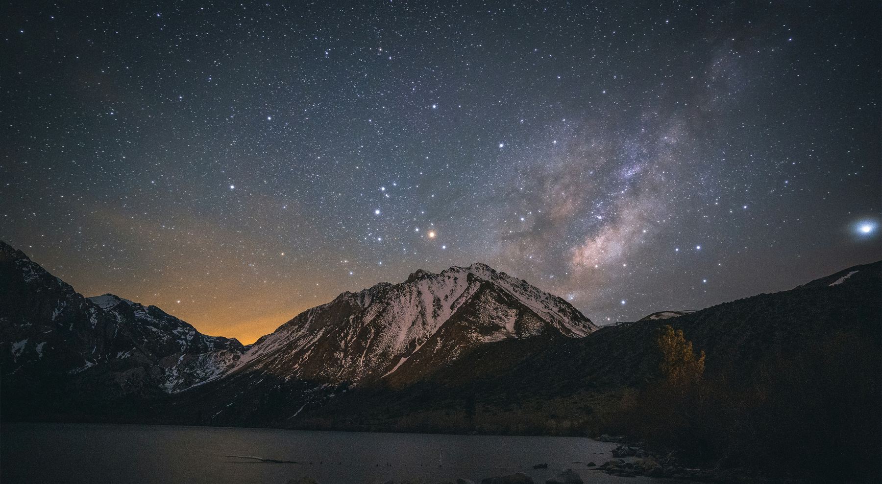 A starry night sky and the Milky Way above snow-capped mountains and a lake.