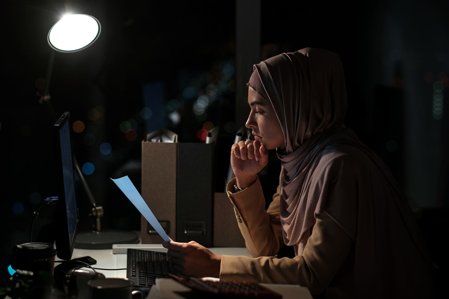 A woman in a hijab working late at a desk under a lamp, reviewing a paper document beside her computer.